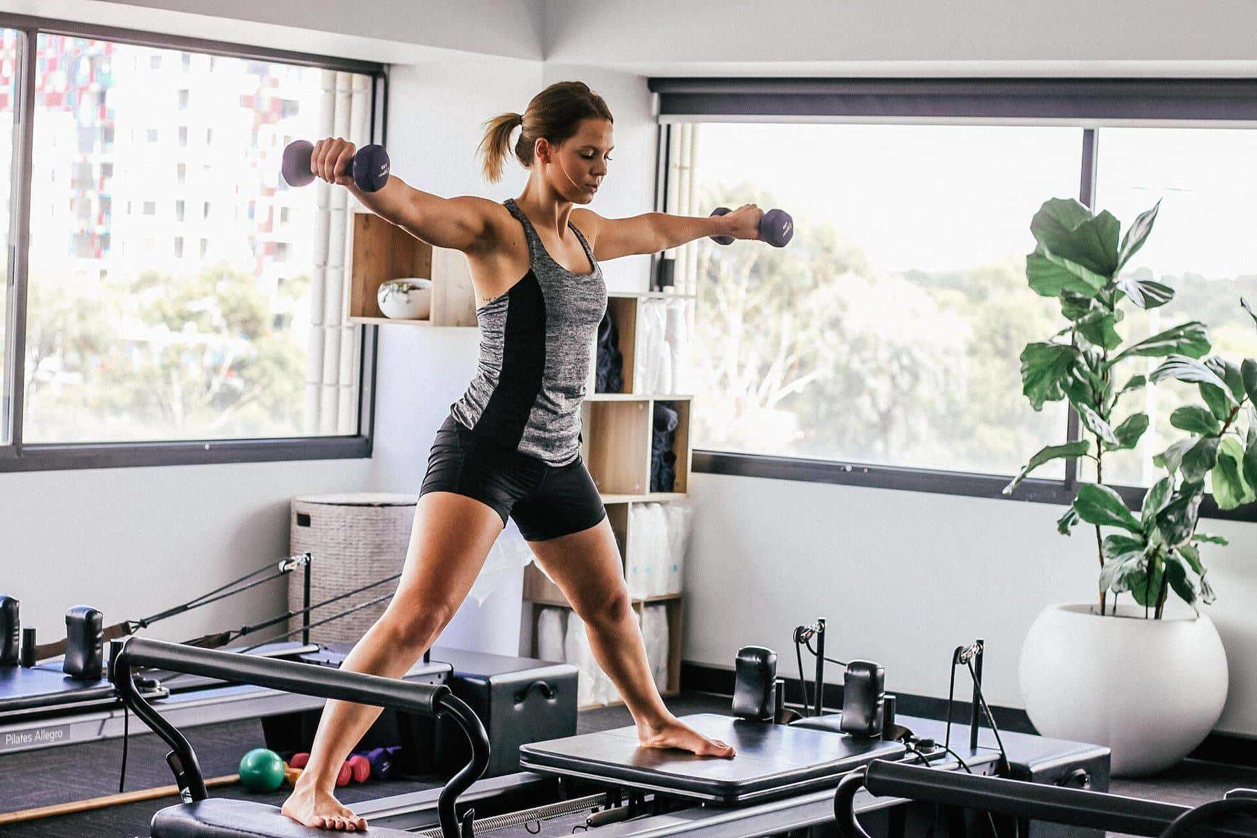 A woman standing on a Pilates reformer holding hand weights in a bright studio