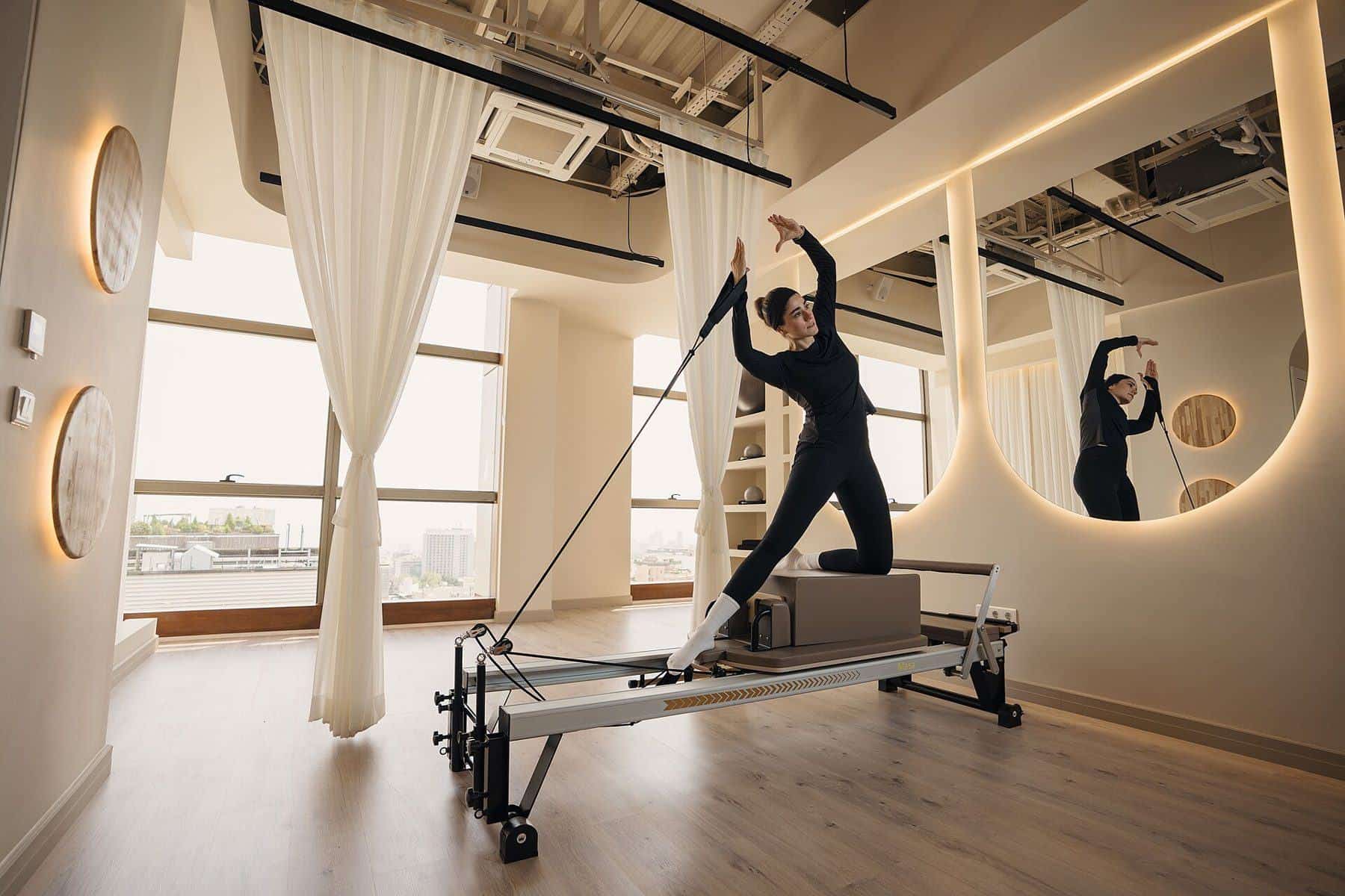 A woman standing on a reformer using the straps in a mirrored Pilates studio